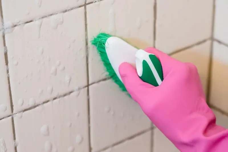 Gloved hand scrubbing bathroom tile grout with a brush, the first step of bathroom mold removal in Cary, NC
